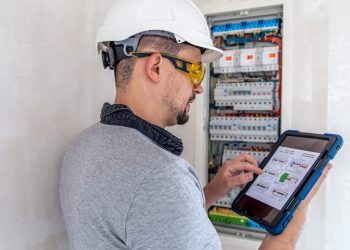 Man, an electrical technician working in a switchboard with fuses. Installation and connection of electrical equipment. Professional uses a tablet.